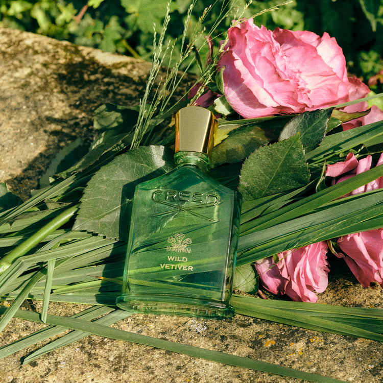 Green Creed perfume bottle on a stone surface with pink roses and green leaves.