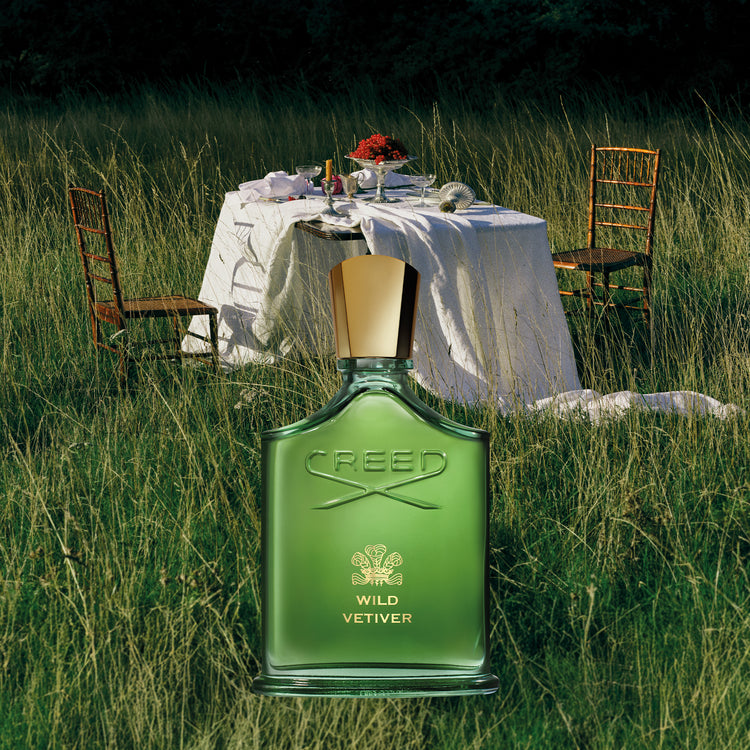 Green Creed Wild Vetiver perfume bottle in a grassy outdoor setting with a table and chairs.