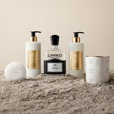 Collection of Creed beauty products including a bottle, two pump bottles, a candle, and a soap on a sandy surface.