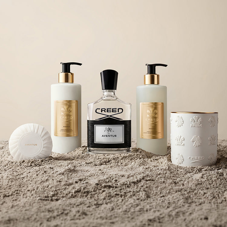 Collection of Creed beauty products including a bottle, two pump bottles, a candle, and a soap on a sandy surface.