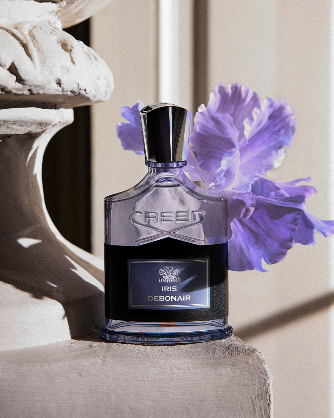 A bottle of Creed Iris Debonair perfume stands on a stone pedestal, with a large purple iris flower behind it and a sculptural stone form to the left. The bottle is clear with a black lower section and a reflective silver cap.