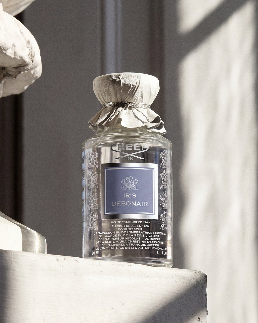 A bottle of Creed Iris Debonair eau de parfum with a clear glass body, silver foil neck wrap, and a lavender-blue label, resting on a white stone ledge in natural sunlight.