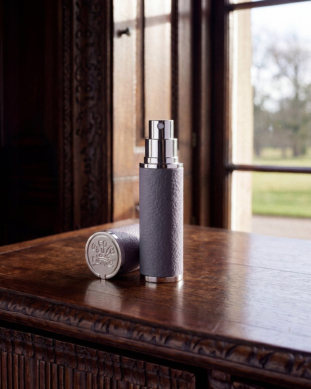 A cylindrical perfume bottle with a textured gray body and a shiny silver spray nozzle stands on a polished wooden table, with its matching cap placed beside it. A window in the background shows a blurred outdoor scene.
