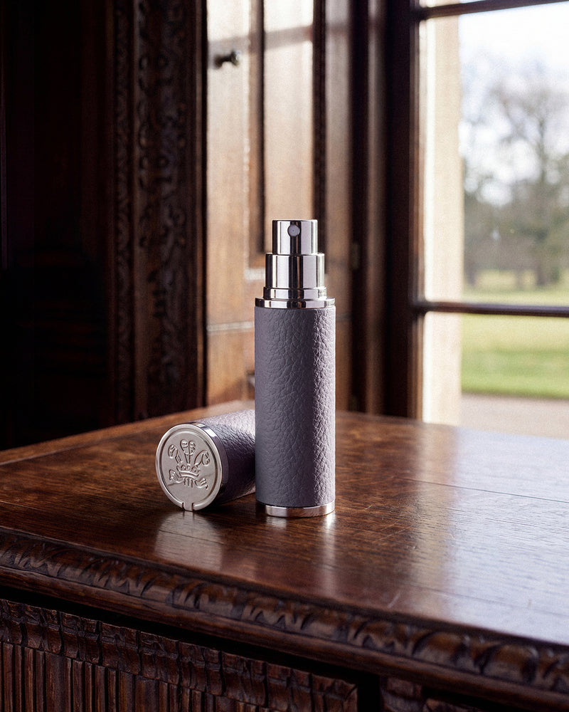 A cylindrical perfume bottle with a textured gray body and a shiny silver spray nozzle stands on a polished wooden table, with its matching cap placed beside it. A window in the background shows a blurred outdoor scene.