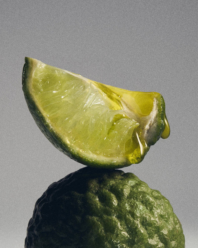 A close-up studio shot of a lime wedge resting on top of a whole lime against a smooth gray background.