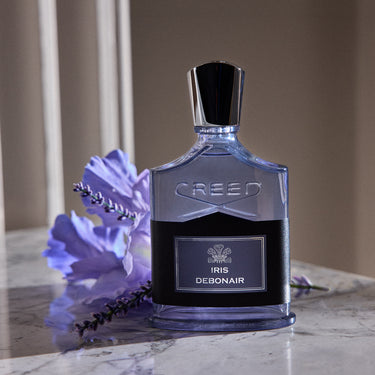 A bottle of Creed Iris Debonair perfume sits on a marble surface next to purple flowers. The bottle is clear glass with a black cap and a black label featuring the silver Creed logo and 'IRIS DEBONAIR' text.