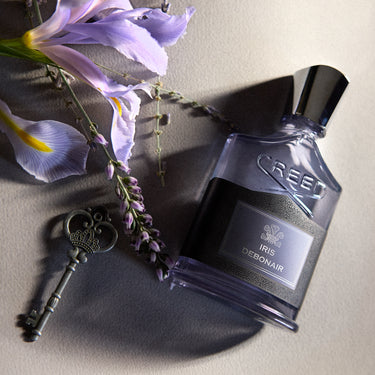 Creed Iris Debonair perfume bottle with a black label and silver cap, styled with purple irises and a vintage key on a textured gray surface.