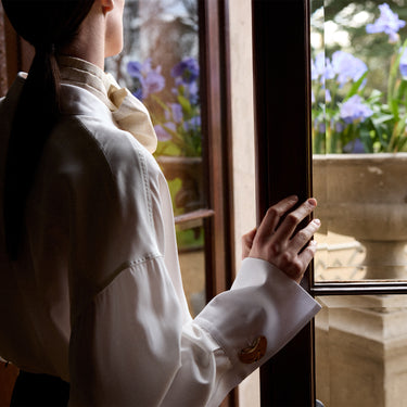 A woman in a white long-sleeve blouse with a cream neck scarf and gold cufflinks looks out of a window, with purple flowers visible outside.