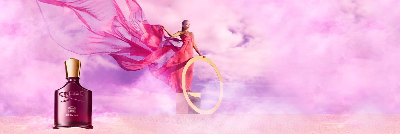 A model with a flowy pinkish dress in a purple clouds with a large gold wing. In the forfront there is a Carmina 75ml bottle, to the left