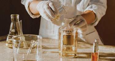 A person wearing white gloves and a white lab coat pours liquid from a small glass container into a clear bottle labeled CREED, set on a tabletop with glassware and a small perfume spray nearby.