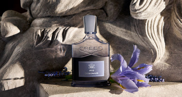Creed 1915 Lebanon fragrance bottle displayed on a stone surface with a purple iris flower beside it. The bottle is dark glass with a silver-toned cap and front label, set against a sculptural stone background.