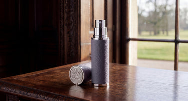 A cylindrical perfume bottle with a textured gray body and silver metallic collar, standing on a polished wooden table next to its matching cap, with a window showing a blurred outdoor garden in the background.