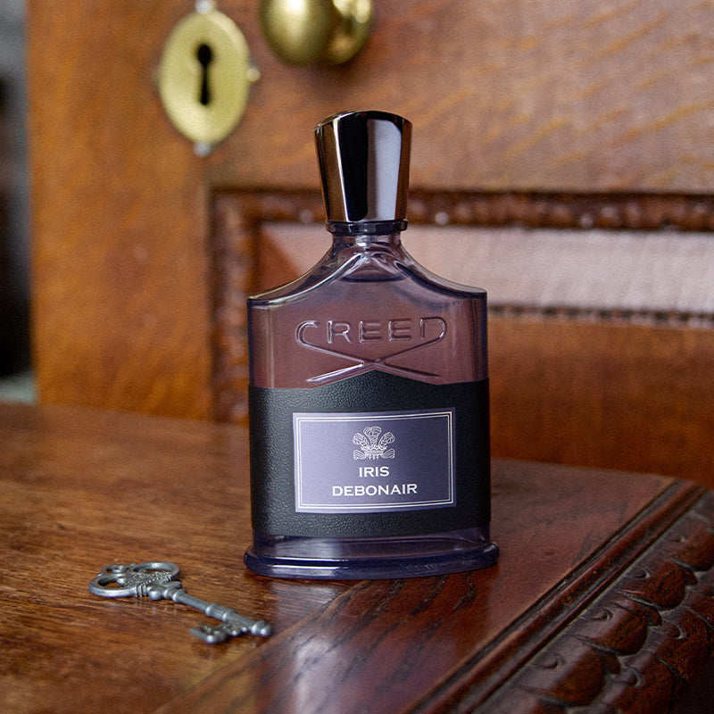 A bottle of Creed Iris Debonair perfume stands on a polished wooden table, with a silver key nearby and a warm wooden cabinet background featuring a brass keyhole.
