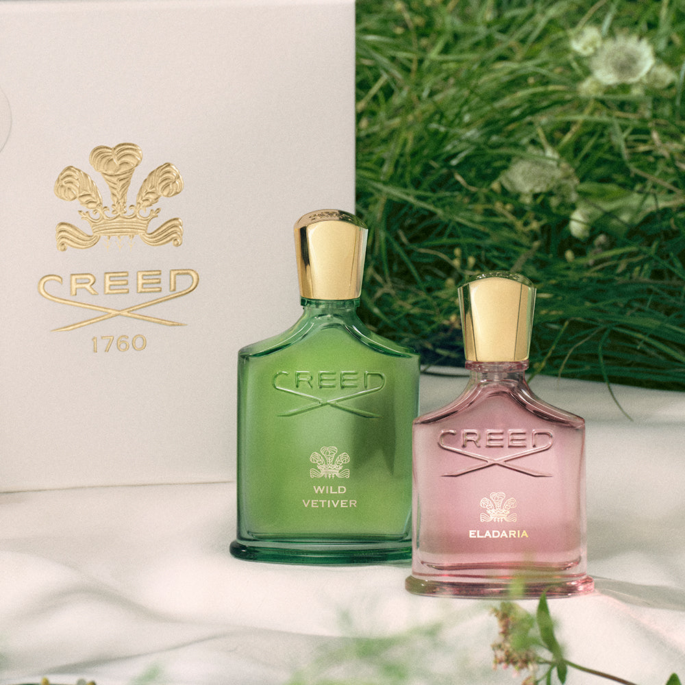 Two Creed perfume bottles, one green and one pink, with a white box featuring the Creed logo in the background.