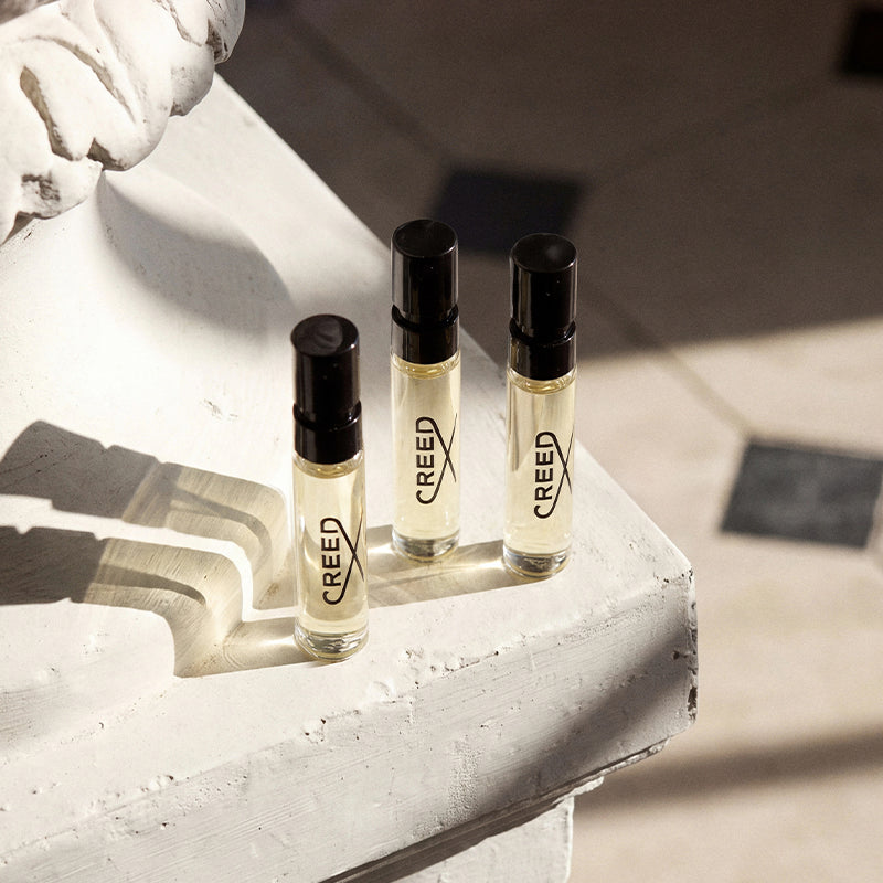 Three small clear glass perfume or fragrance bottles with black caps and visible CREED branding, arranged on a white painted stone ledge in bright sunlight.