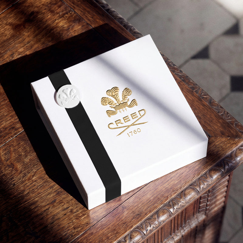 A white Creed gift box with a black vertical stripe, gold crest logo, and a silver circular seal, resting on a dark wooden table in warm sunlight.