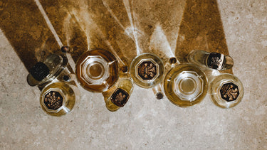 A group of clear glass bottles with gold-toned liquid and black caps, arranged on a textured beige stone surface under direct sunlight.