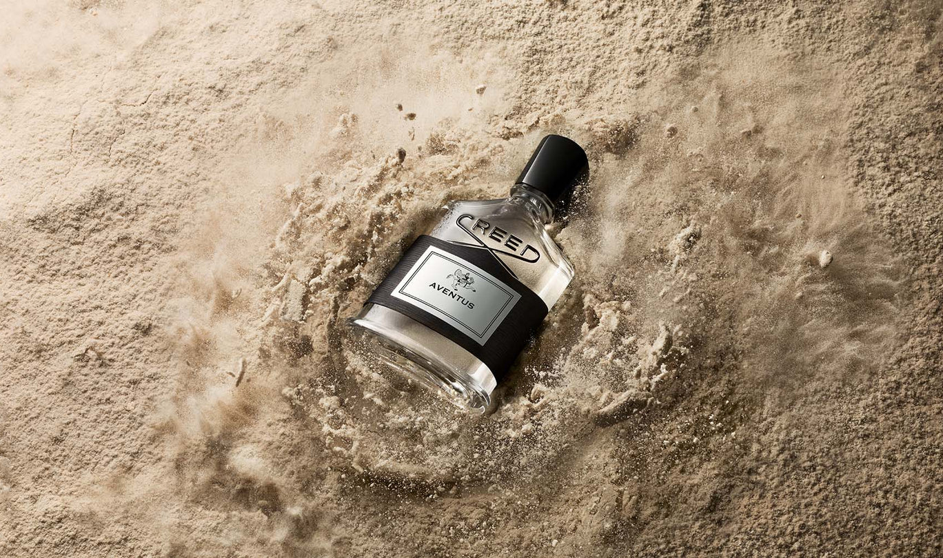 A bottle of Creed Aventus perfume lies partially submerged in wet sand, with water and sand splashing around it. The bottle is clear glass with a black cap and a black-and-silver label.