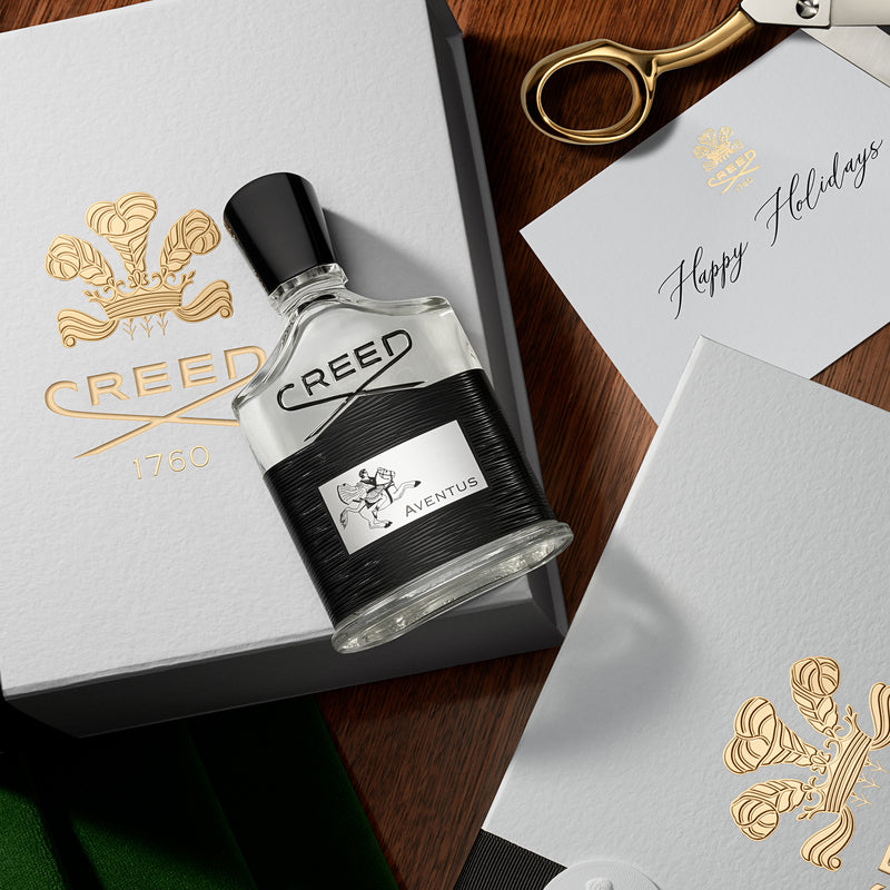 Creed Aventus perfume bottle on a wooden surface with a box and 'Happy Holidays' card.