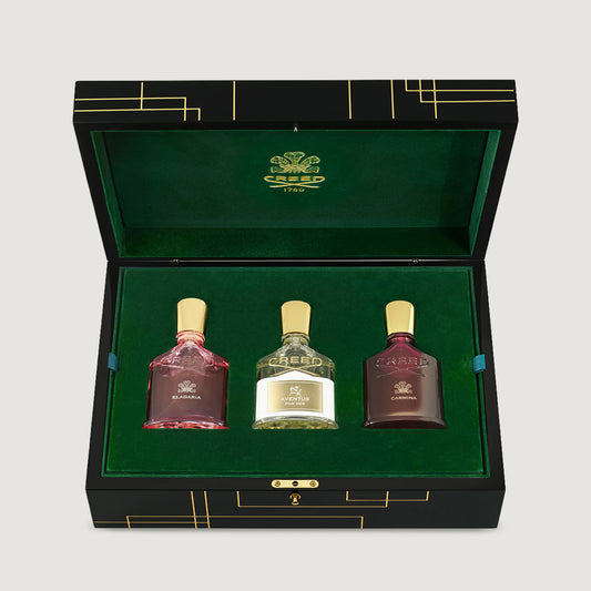 An open black presentation box with a green velvet interior displaying three small bottles from the brand CREED, set against a light gray background.