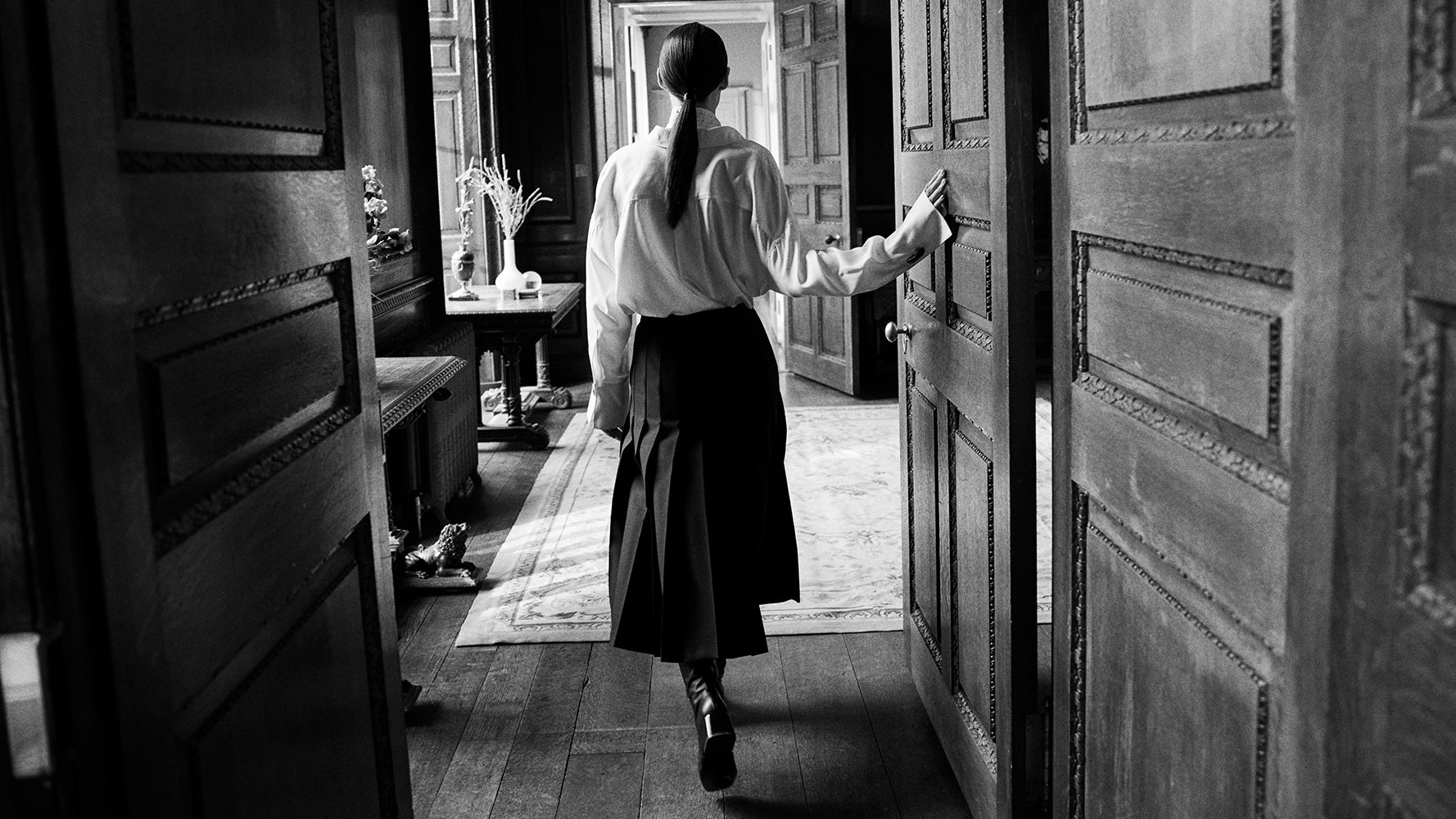 A black and white photograph of a woman seen from behind, wearing a white blouse and a dark skirt, walking through an open doorway into a dimly lit room.