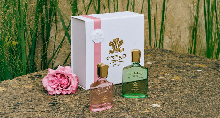 Two Creed perfume bottles with a box on a stone surface with grass in the background